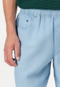 Pantalons bleu clair en tissu doux, dotés d'une taille élastique, de poches latérales et de détails en métal doré.