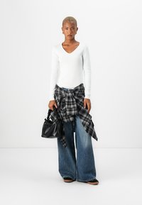 Hvit langermet v-hals genser, oversized blå denimjeans, knyttet svart og hvit rutete skjorte, svart veske i teksturert materiale, avslappede sko.