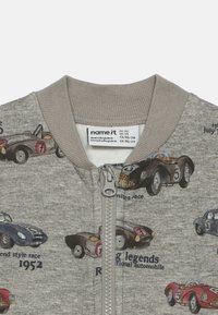 Γκρι φούτερ με φερμουάρ, διακοσμημένο με εικονογραφήσεις vintage αγωνιστικών αυτοκινήτων και κείμενα όπως "1952" και "θρύλοι του δρόμου". Η γιακάς είναι φτιαγμένη από πλέκτο ύφασμα.