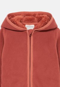 Veste en polaire de couleur terracotta, dotée d'une fermeture éclair, d'une capuche et d'une doublure douce et texturée. Conçue pour les nourrissons, avec un confort douillet.