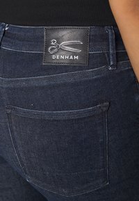 Vaqueros de denim azul oscuro Denham con una etiqueta de marca de cuero negro que presenta tijeras. Tiene costuras visibles y un bolsillo trasero.