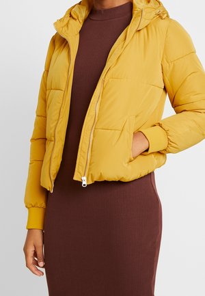 Mujer con chaqueta acolchada de color mostaza sobre un vestido midi marrón acanalado, con una mano en el bolsillo de la chaqueta y la otra relajada al lado.