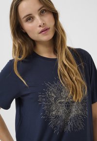 T-shirt bleu marine en tissu doux, avec un design graphique central doré présentant des motifs complexes et des lignes rayonnantes.