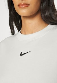 Sudadera gris claro de tela suave con cuello redondo acanalado. Presenta un pequeño logo negro de Nike en la parte superior izquierda.
