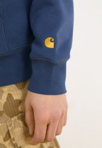 Mørkeblå sweatshirt med ribbestrikkede mansjetter; har en liten gul logo på venstre erme. Brukt over et mönstrete beige skjørt.