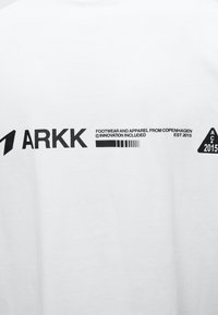 Λευκό ύφασμα με μαύρο κείμενο που λέει ARKK, "Footwear and apparel from Copenhagen, innovation included, est 2015," και ένα τρίγωνο λογότυπο με "AC 2015."