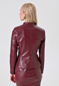 Veste en simili cuir bordeaux au design ajusté, avec un col haut et des détails de couture. Texture lisse, contrastant avec une jupe assortie.