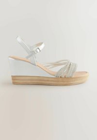 Hvide kilehæle med en sølvrem og perledetaljer over foden. Har en tekstureret beige platform og en sikker ankelrem.
