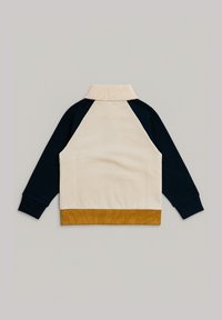 Veste à manches longues avec des manches bleu marine, un corps beige, un col côtelé et un ourlet côtelé jaune moutarde, posée à plat sur une surface grise.