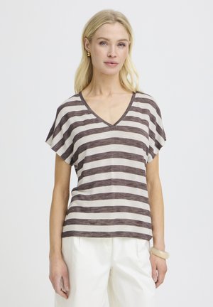 Blondína v hnedoružovobielom tričku s výstrihom do V a vodorovnými pruhmi, bielych nohaviciach, zlatých náušniciach a béžovom náramku stojaca na bielom pozadí.