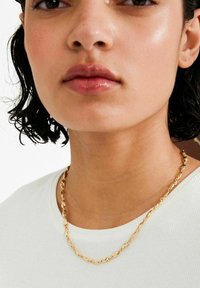 Kuldne kett-kaelakee keerdunud linkidega, läikiva viimistlusega. Kanditud valge soonilise särgi üle mudelil, kellel on lainelised juuksed.