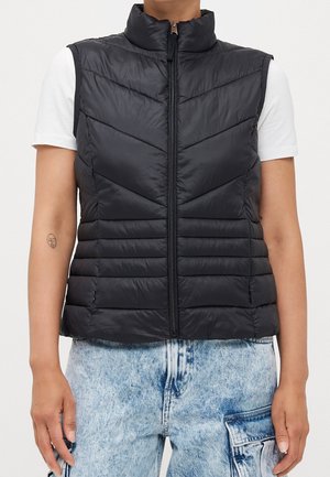 Czarna pikowana kamizelka bez rękawów z zamkiem z przodu, noszona na białej koszuli z krótkim rękawem oraz jasnoniebieskich jeansach cargo z efektem acid wash.