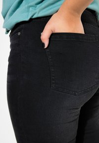 Jeans de mezclilla negra mostrados a la altura de la cadera, con un bolsillo trasero y una mano insertada. El material parece suave con un ligero brillo.