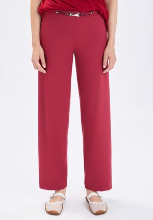 Donna che indossa pantaloni rossi a gamba larga a vita alta con cintura abbinata e scarpe basse bianche, in piedi contro uno sfondo neutro.