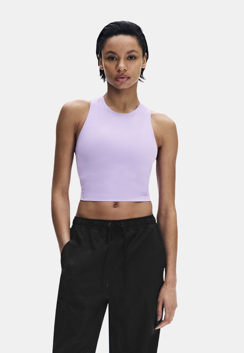 Jeune femme portant un crop top lavande sans manches et un pantalon noir à cordon, debout les mains dans les poches devant un fond uni.
