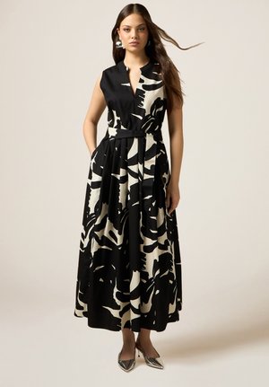 Mujer con vestido midi sin mangas con estampado abstracto en blanco y negro, escote en V y tacones metálicos puntiagudos, de pie frente a un fondo liso.