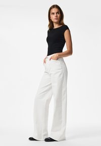 Femme debout les mains dans les poches portant un top noir ajusté à manches courtes et un pantalon large blanc taille haute avec des chaussures noires.