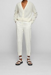 Jasnokremowy, dziergany kardigan z dekoltem w serek i pięcioma guzikami, zestawiony z dopasowanymi, białymi spodniami z mankietami oraz sandałami na czarnych paskach.