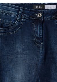 Jeans de mezclilla azul oscuro con un diseño clásico, que cuentan con un bolsillo frontal y un cierre de botón plateado; detalles de costura visibles en todo lo largo.