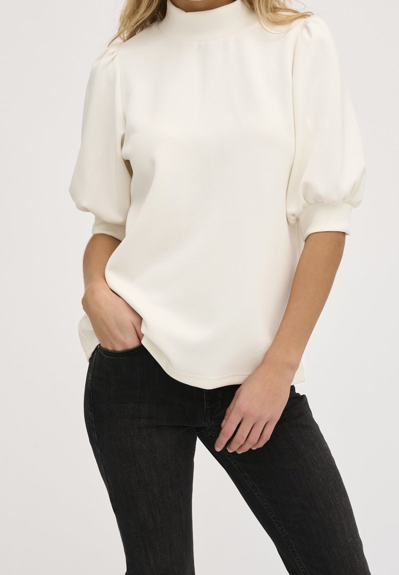 Haut blanc à col haut avec manches bouffantes, en tissu lisse. Associé à un jean noir ajusté. Design simple avec peu de détails.