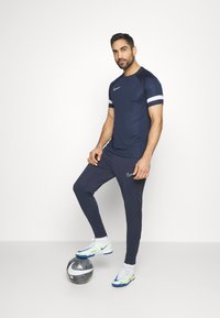 Mand i navy atletisk tøj med hvide striber på ærmerne står med den ene fod på en sort og hvid Nike fodbold foran en hvid baggrund.