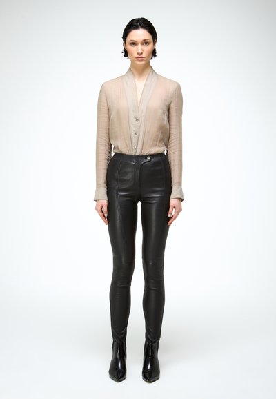 Jonge vrouw staand, vooruit kijkend, met een doorschijnende beige blouse met knopen, zwarte leren broek met hoge taille en zwarte enkellaarzen.