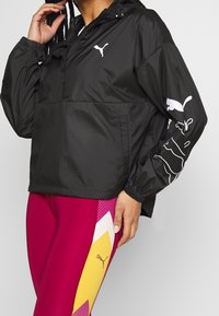 Svart vindjacka med långa ärmar, med en framficka och vitt Puma-logga. Bärs med lysande rosa och gula leggings.