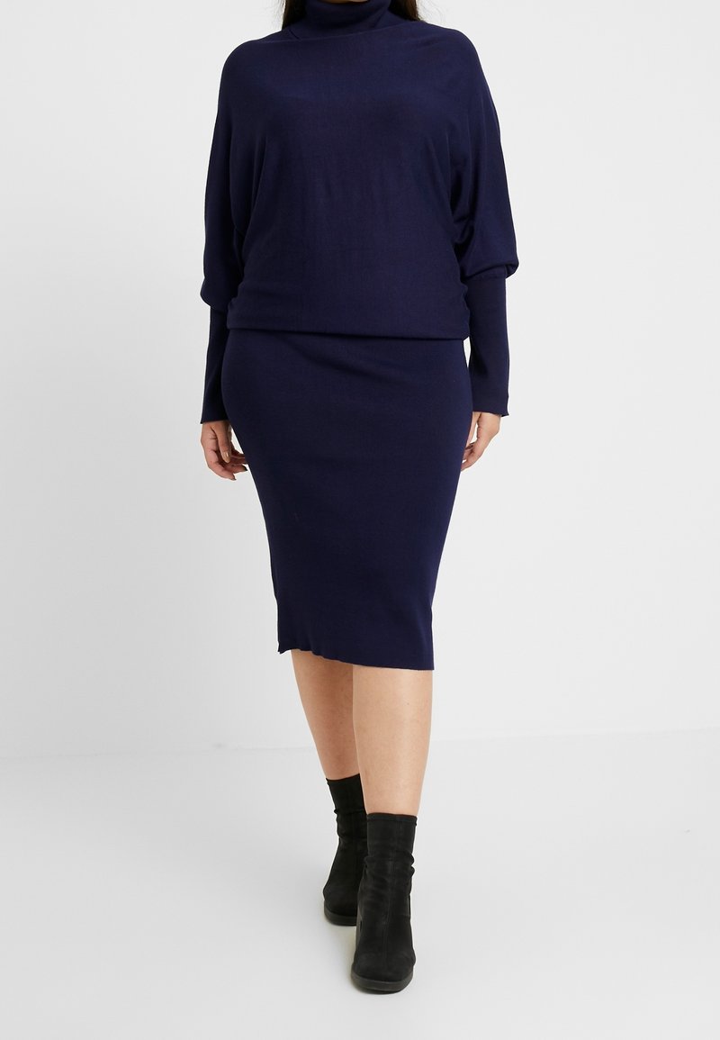 Femme portant un haut à col roulé à manches longues bleu marine et une jupe crayon assortie longueur genou, avec des bottines noires, sur fond blanc.