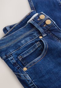 Sötétkék farmernadrág összehajtva, elöl zseb, övhurkok, dupla arany gombok, szegecsek és részletes narancssárga varrások fehér háttéren.