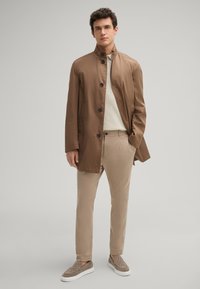 Bruine lichtgewicht jas met knopen, beige sweater, lichtbruine broeken en grijze instapschoenen met een textuurpatroon.