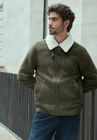 Chaqueta verde oliva con un suave collar de borreguillo blanco, que presenta un cierre de cremallera, bolsillos laterales y una textura de ante suave.
