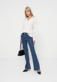 Blusa branca de renda com um corte relaxado e colarinho, combinada com jeans flare azuis com riscas; modelo carrega uma pequena mala preta.