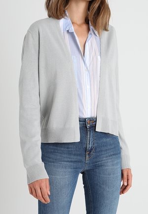 Kobieta ubrana w jasnoszary kardigan z otwartym przodem na niebiesko-białej pasiastym, zapinanym na guziki koszuli i niebieskich dżinsach, na jednolitym tle.