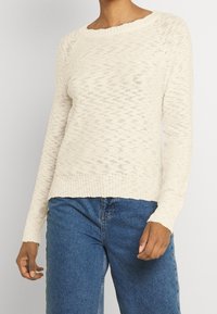 Ljusbeige stickad tröja med en texturerad design, vid halsringning och ribbade ärmslut; matchas med blå denimjeans.