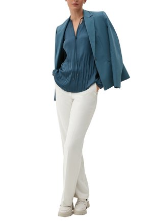 Blusa plissettata color turchese con cordoncino, pantaloni bianchi a gamba dritta e scarpe beige slip-on. La giacca è di un solido color turchese con una vestibilità su misura.