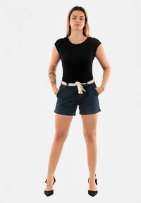 Zwart nauwsluitend topje gecombineerd met donkerblauwe korte broek met een wit patroonriem. Model draagt zwarte hakken en poseert met handen in de zakken.