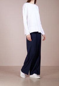 Žena oblečená v volné bílé dlouhé tričko s dlouhým rukávem, tmavě modrých širokých kalhotách a bílých teniskách, stojící na béžové podlaze proti světle hnědé zdi.