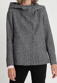 Šedý kabát v herringbone vzoru s kapucí, zapínáním na knoflíky vpředu a žebrovanými růžovými manžetami. Vyroben z texturované tkaniny, která poskytuje strukturovaný siluetu.