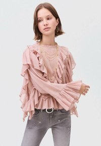 Blusa rosa chiaro con volant, scollo rotondo e maniche ampie, abbinata a jeans grigi con una fibbia rotonda a contrasto.