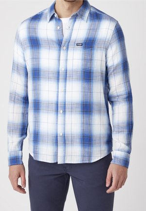 Mężczyzna w niebiesko-białej kraciastej koszuli na guziki z długim rękawem, na białej podkoszulce i ciemnych spodniach, stojący na jednolitym tle.