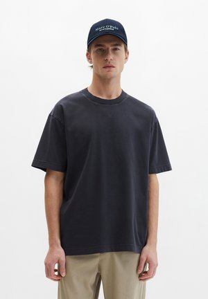 Młody mężczyzna w granatowej czapce Marc O'Polo, ciemnej, oversize'owej koszulce i beżowych spodniach, stojący na jednolitym białym tle.