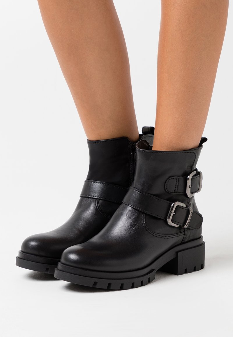 Bottes noires en cuir à cheville avec semelles épaisses et boucles argentées portées sur des jambes nues sur fond blanc.