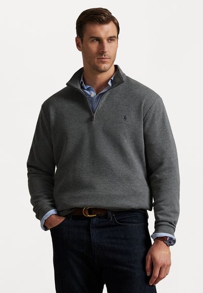 Homme portant un pull à col zippé gris foncé par-dessus une chemise col bleu clair, un jean noir, une ceinture marron et une montre au poignet, debout avec une main dans la poche.