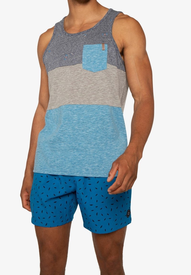 Homme portant un débardeur sans manches à blocs de couleurs avec une poche poitrine et un short de bain bleu orné de petits motifs noirs.