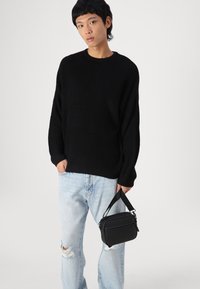 Pull noir à côtes, jean bleu clair déchiré avec des trous, et un petit sac à bandoulière noir avec fermeture éclair et bandoulière ajustable.
