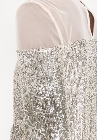 Silbernes Paillettenoberteil aus transparentem, leichtem Material mit lockerem Schnitt und schulterfreiem Design, das schimmernde Muster zeigt.