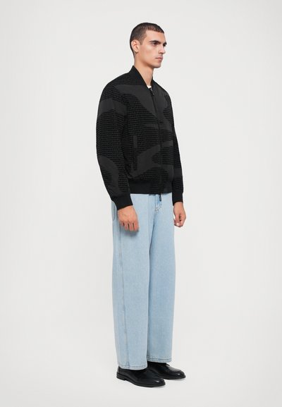 Black bomber jacket with a textured design, featuring a zip front and ribbed cuffs. Worn with light blue wide-leg jeans and black shoes.