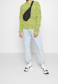 Lime green knitted sweater, vaaleansiniset löysät farkut ja valkoiset lenkkarit, joissa on siniset yksityiskohdat. Musta olkalaukku kantaa vasemmalla puolella.