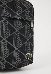 Primo piano di una borsa in tessuto nero con motivo geometrico grigio, zip argentata e un piccolo logo a forma di coccodrillo verde nell'angolo inferiore.