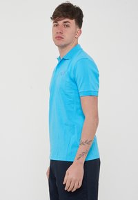 Polo shirt azzurro chiaro con colletto, maniche corte e un logo sottile sul lato sinistro del petto. Tessuto in cotone, texture liscia, design aderente.